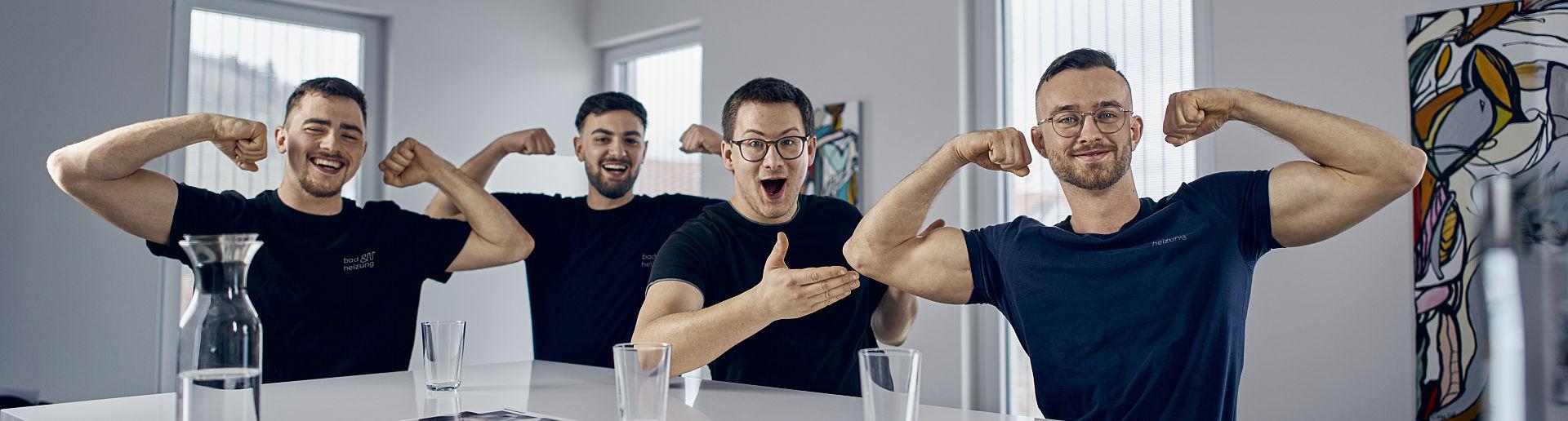 Junge Männer zeigen ihre Muskeln in einem hellen Raum, während ihre Kollegen lachend applaudieren.