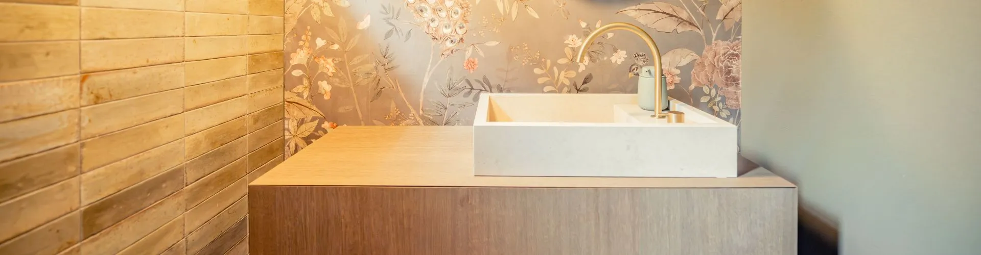 Luxuriöses Aufsatzwaschbecken mit goldener Armatur vor Motivtapete mit Blumen und Vogelmotiven.