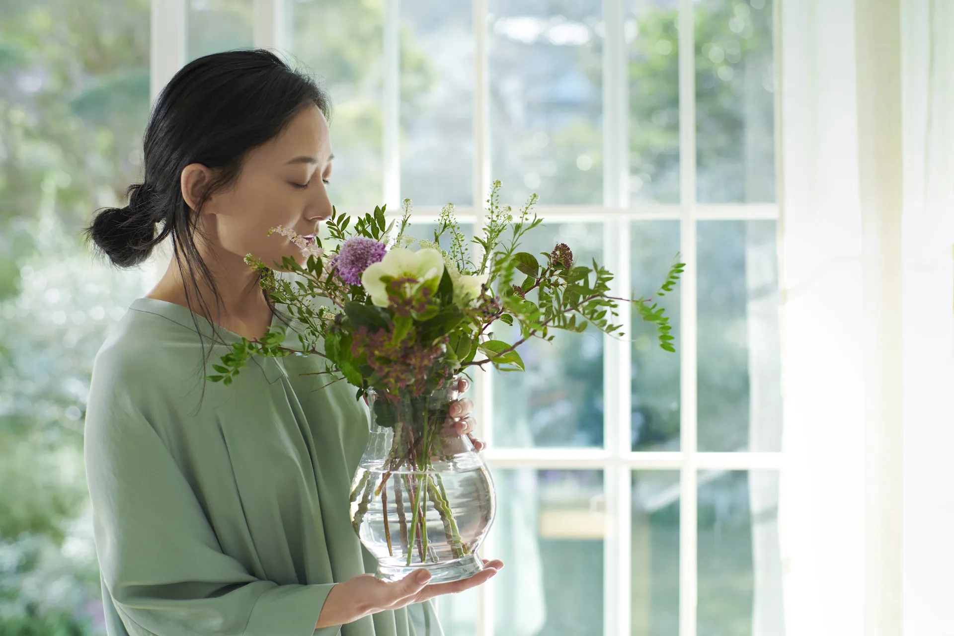 Japanische Frau steht vor einem großen Fenster und hält eine Vase mit wohlduftenden Blumen in der Hand.
