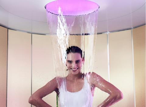 Eine Frau steht unter einem breiten Wasserstrahl in einer luxuriösen Dusche, beleuchtet von lila Licht, und genießt das entspannende Spa-Erlebnis.