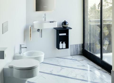 Ein modernes Badezimmer mit weißen Wänden und einem Boden aus hellem Marmor. Links sind eine schwebende Toilette und ein Bidet zu sehen, rechts davon ein rundes Waschbecken mit einem schwarzen Regal darunter. Durch die große Glasfront im Hintergrund fällt viel natürliches Licht ins Zimmer.