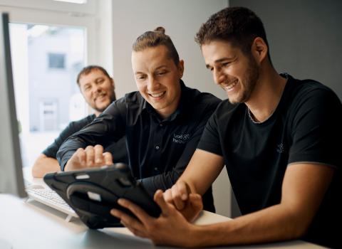 Drei Kollegen stehen an einem Schreibtisch und arbeiten gemeinsam an einem Tablet, das von einem der Männer gehalten wird. 