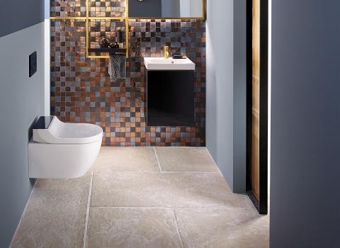 Elegantes Bad mit Wand-WC Geberit AquaClean Tuma, Mosaikfliesen und einem schmalen, goldgerahmten Spiegel. Dezente Beleuchtung schafft eine warme Atmosphäre.