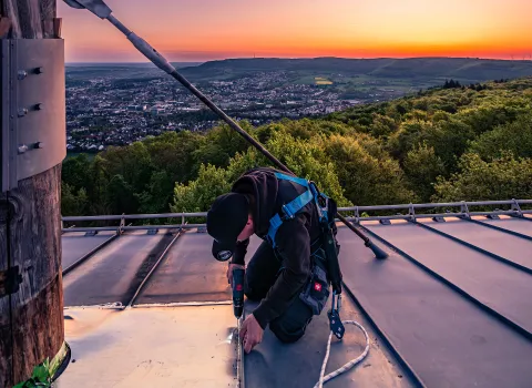 Ein Handwerker in Sicherheitsausrüstung montiert ein Metallbauteil auf einem Dach bei Sonnenuntergang.bäumöl