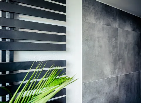 Ein moderner Designheizkörper in horizontalen, schwarzen Lamellen ziert die Wand eines Badezimmers. Daneben sind großflächige Fliesen in grauer Betonoptik zu sehen.