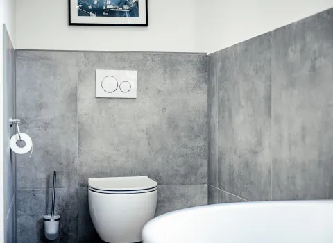 Ein modernes WC mit Wandmontage ist vor großformatigen Fliesen in grauer Betonoptik installiert. Darüber befindet sich eine weiße Betätigungsplatte mit zwei Spültasten.