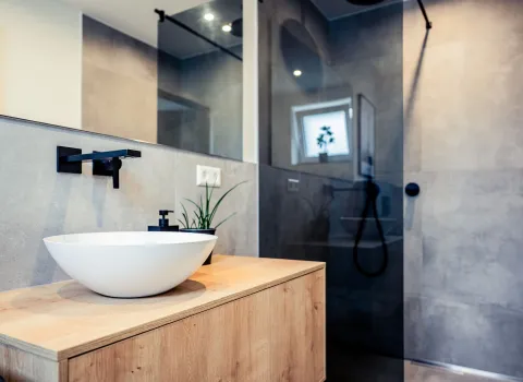Das stilvolle Bad kombiniert natürliche Holzoberflächen mit industriell wirkenden Betonfliesen. Das weiße Aufsatzwaschbecken steht im Kontrast zur mattschwarzen Armatur, während die bodengleiche Dusche mit Glaswand und Regendusche für Offenheit sorgt.