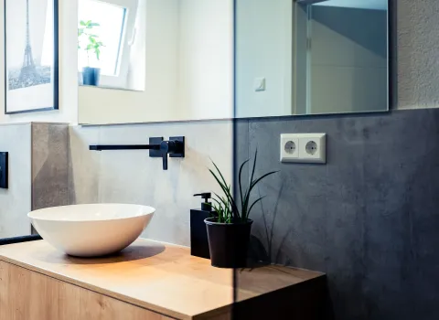 Das moderne Gästebad besticht durch klare Linien, ein rundes Keramikwaschbecken und eine dunkle Wandarmatur im Industrial-Stil. Der Holzwaschtisch verleiht dem Raum Wärme, während graue Fliesen einen modernen Akzent setzen.