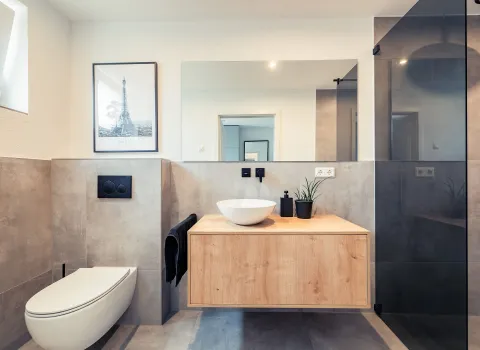 Frontalansicht des Gästebades mit großem, wandhängendem WC und rechts daneben der Waschplatz mit Holzunterschrank, Keramik-Aufsatzbecken und großen rechteckigen Spiegel.