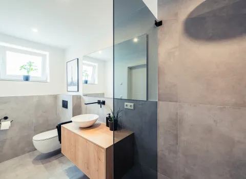 Ein helles Gästebad mit wandhängendem WC, rundem Aufsatzwaschbecken auf einem schwebenden Holzwaschtisch und großer Spiegelwand.