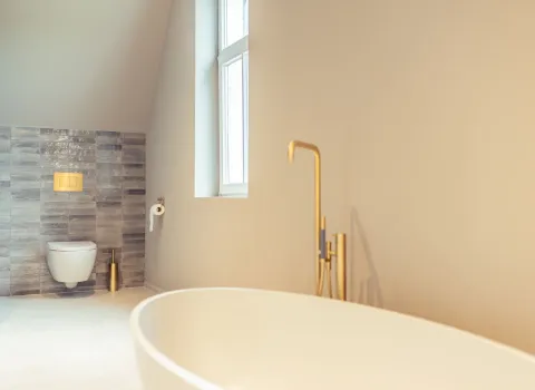Im Vordergrund steht eine weiße, freistehende Badewanne mit goldener Standarmatur. Rechts fällt Tageslicht durch ein Fenster, links ist eine Wand mit grauen Fliesen und wandhängendem WC zu sehen.