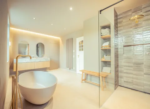 In einem hellen Raum steht eine freistehende Wanne mit goldener Armatur, dahinter ein stilvoller Doppelwaschtisch mit Spiegeln. Die begehbare Dusche mit Glasabtrennung ist mit glänzenden, grauen Fliesen verkleidet.