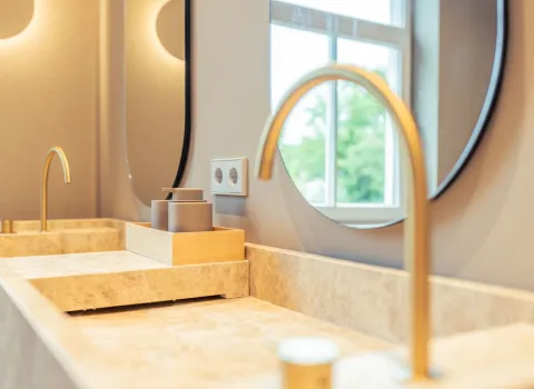 Zwei goldene Armaturen ragen über ein Waschtisch-Ensemble aus beigem Naturstein. Im Hintergrund spiegeln sich Fenster und Lichtquellen in den abgerundeten Spiegeln.