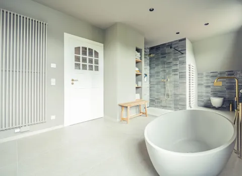 Dieses helle Premium-Bad zeigt eine stilvolle Kombination aus skandinavischer Schlichtheit und modernem Luxus. Eine frei stehende Badewanne mit goldener Armatur steht im Vordergrund, während eine begehbare Dusche mit grauen Wandfliesen den Hintergrund bildet.