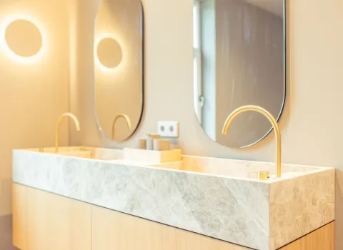 Der Waschtisch mit Marmoroberfläche und zwei filigranen goldenen Armaturen steht vor einer hellen Wand mit runden Wandlichtern.