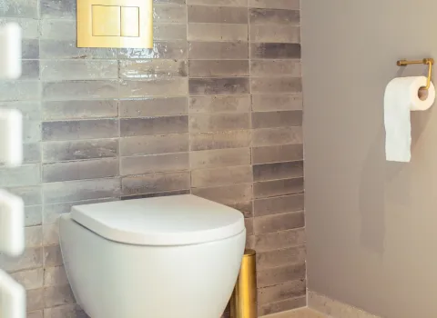 Ein wandhängendes WC vor einer Wand mit glänzenden, grauen Fliesen. Darüber ein rechteckiger Spülknopf in Goldoptik, daneben ein goldener Halter für die Toilettenbürste.