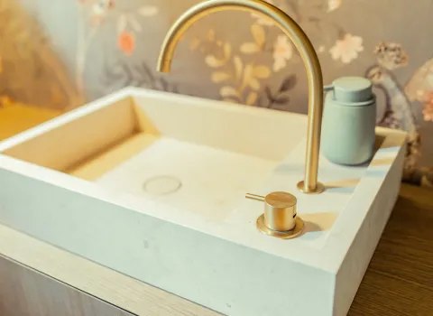 Ein rechteckiges Aufsatzwaschbecken aus weißem Stein steht auf einem Holzunterschrank und wird von einer gebogenen, goldfarbenen Armatur ergänzt.