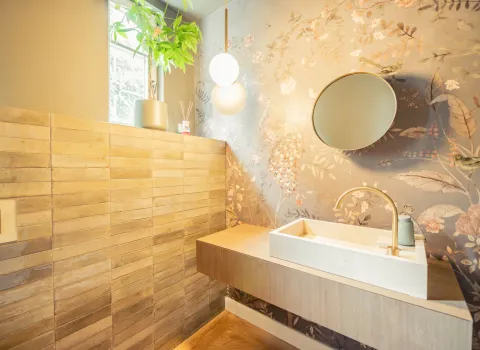 Ein modernes Badezimmer mit floraler Motivtapete, die durch das einfallende Licht vom Fenster besonders zur Geltung kommt. Das goldfarbene Wasserhahn-Design ergänzt das elegante Waschbecken auf dem schwebenden Waschtisch. Eine minimalistische Hängelampe sorgt zusätzlich für warmes Licht. Die linke Wand ist mit strukturierten Holzfliesen verkleidet.
