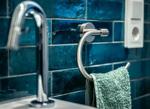 Im Vordergrund ist eine moderne, gebogene Armatur aus Chrom über einem weißen Waschbecken zu sehen. Daneben hängt ein grünes Handtuch an einem runden Handtuchhalter aus Edelstahl, der an einer Wand mit glänzenden, dunkelblauen Fliesen befestigt ist.