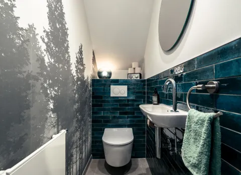 Frontaler Blick ins fensterlose, schlauchförmige Gäste WC mit Motivtapete und blauen Fliesen.