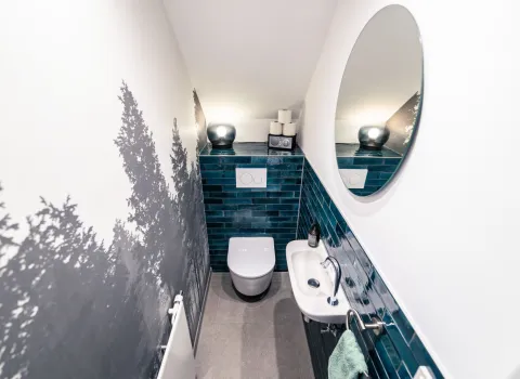 Schlauchförmiges enges Gäste-WC mit Tapete mit Waldmotiv, blauen Fliesen und runden Spiegel über dem schmalen Waschbecken..