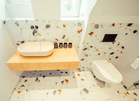 Helles Bad mit Terrazzo-Wand und Boden in bunten Farbsplittern. Holzplatte mit weißem Waschbecken und wandhängendem WC sorgen für ein luftiges Raumgefühl.