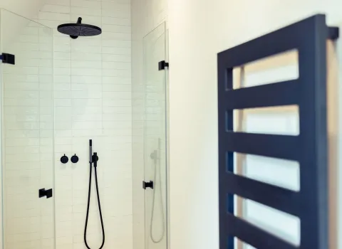 Moderne Dusche mit schwarzer Regenduschkopf-Armatur und klaren Glasabtrennungen vor weiß gefliester Wand. Im Vordergrund ist ein stilvoller, schwarzer Handtuchheizkörper mit rechteckigen Aussparungen zu sehen