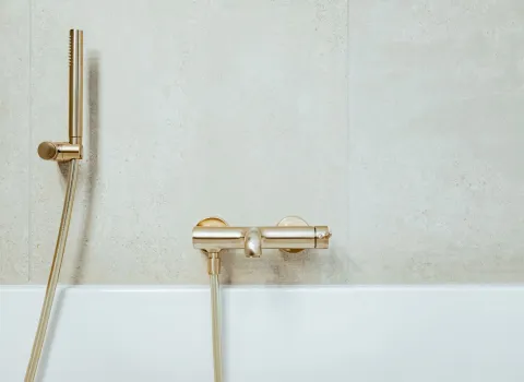 Wandmontierte Badewannenarmatur in Goldoptik vor heller Fliesenwand.