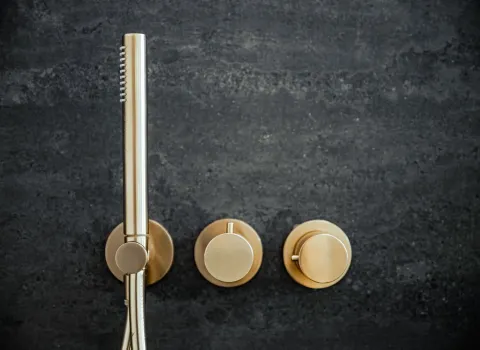 Detailaufnahme einer goldenen Duscharmatur mit Bedienreglern an dunkler Wand.