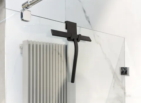 Eine moderne Glasdusche zeigt einen schwarzen Duschabzieher, der mittig an der transparenten Duschwand hängt.