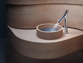 Ein modernes Waschbecken mit einer runden Holzschale, in die ein verchromter Wasserhahn Wasser sprüht. 