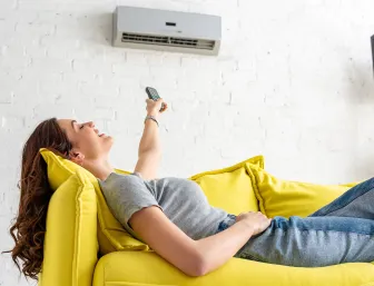 Eine Frau liegt entspannt auf einem leuchtend gelben Sofa und richtet eine Fernbedienung auf die Klimaanlage an der Wand. 