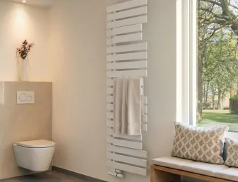Asymmetrischer Handtuchheizkörper in einem hellen Badezimmer. Ein Handtuch hängt an den breiten Streben, daneben ein Fenster mit gemütlicher Sitzbank und Kissen.
