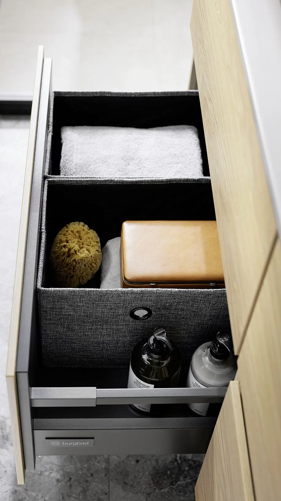 Nahaufnahme einer offenen Schublade von burgbad, die mit einer grauen Stoffbox für Handtücher und Zubehör wie eine Bürste und eine Ledertasche ausgestattet ist.