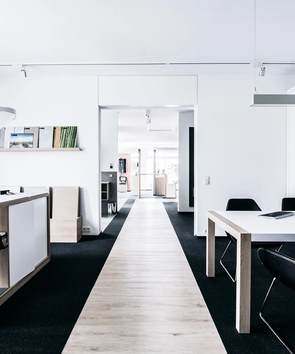 Ein moderner, hell gestalteter Besprechungsraum mit einem weißen Tisch und schwarzen Stühlen vor einer großen Wandnische mit integriertem Bildschirm. 