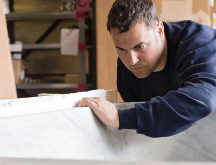 Ein Handwerker inspiziert sorgfältig eine große Badewanne aus Marmor, umgeben von Kartons und Regalen in einer Werkstatt.