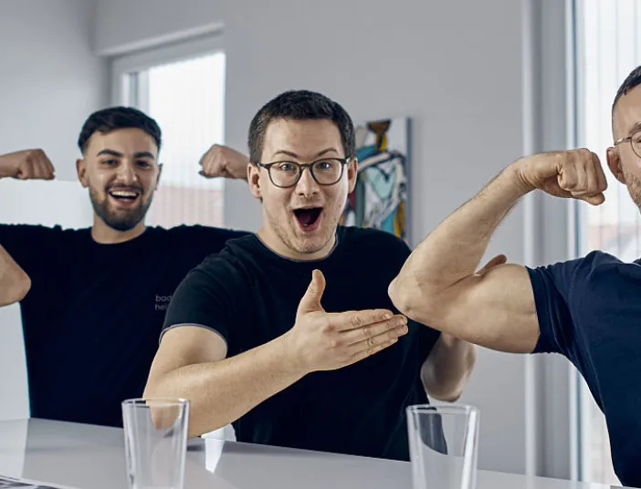 Junge Männer zeigen ihre Muskeln in einem hellen Raum, während ihre Kollegen lachend applaudieren.