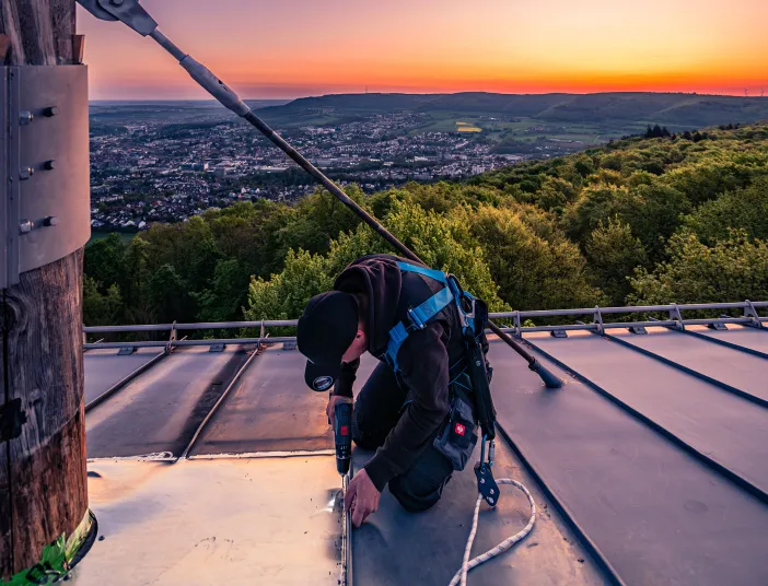 Ein Handwerker in Sicherheitsausrüstung montiert ein Metallbauteil auf einem Dach bei Sonnenuntergang.bäumöl