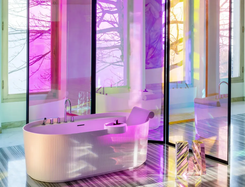 Eine luxuriöse, freistehende Badewanne in einem modernen Badezimmer ist von bodentiefen Fenstern umgeben, die farbiges Licht in Rosa, Gelb und Blau reflektieren. 